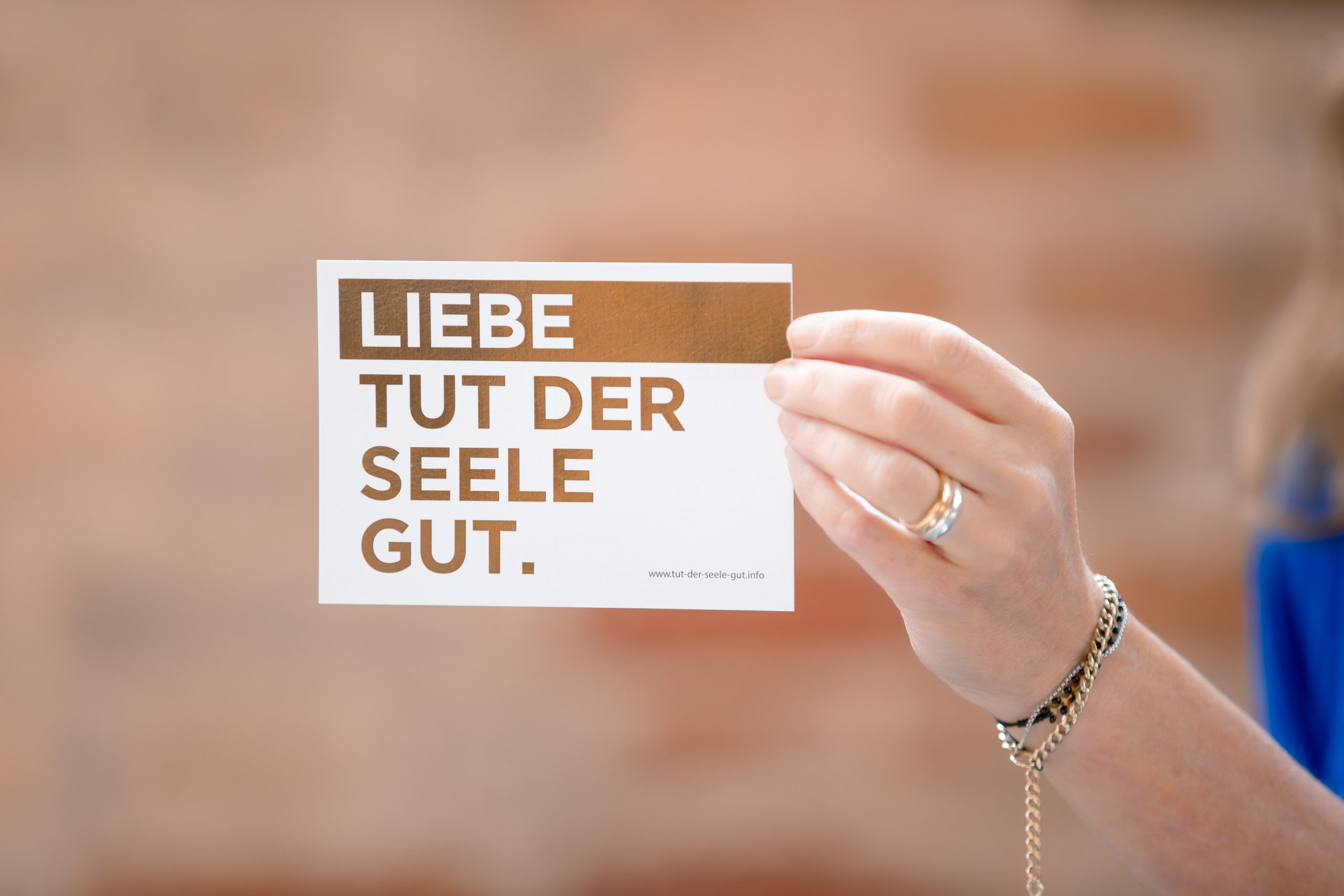 Eine Hand mit Armbändern und einem Ring hält eine Karte mit der Aufschrift „LIEBE TUT DER SEELE GUT“. Der Hintergrund ist in warmen Tönen unscharf.