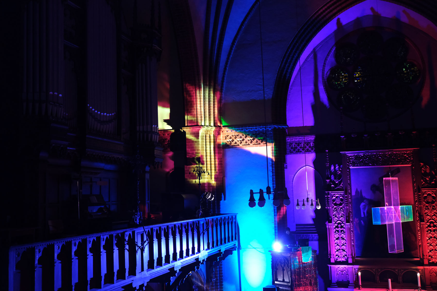 Bunte Lichter erhellen das Innere einer Kirche und werfen leuchtende Muster auf die Wände und den Altar. An der Vorderseite ist ein leuchtendes Kreuz zu sehen, und darüber befinden sich Buntglasfenster. Die Atmosphäre ist dramatisch und kunstvoll.