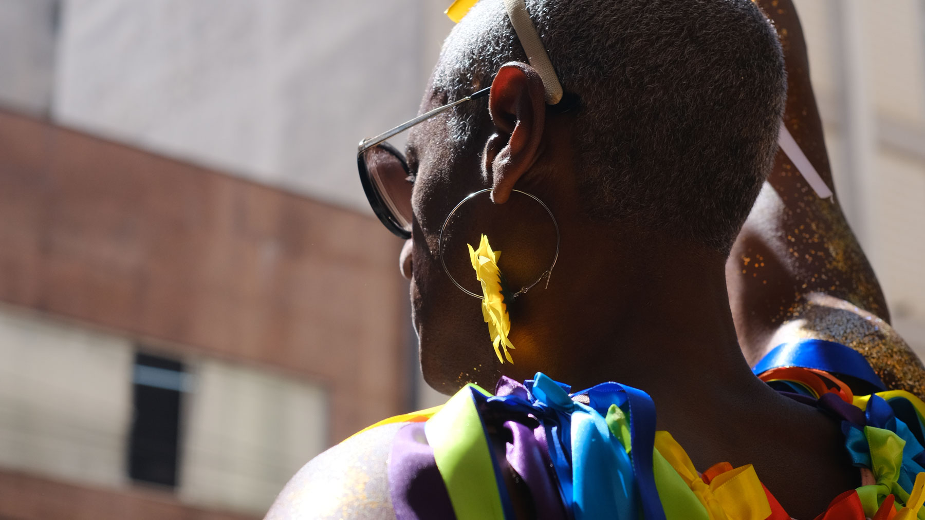 Eine Person mit kurzem Haar und Sonnenbrille trägt einen großen, gelb verzierten Ohrring. Sie hat bunte Bänder über die Schulter drapiert und goldenen Glitzer auf der Haut und steht im Freien im Sonnenlicht.